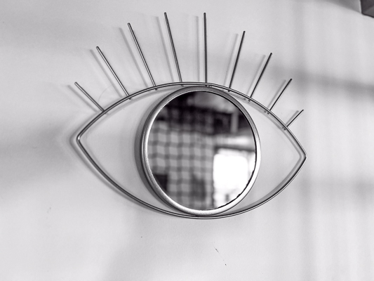 Spiegel in Form eines Auges an weißer Wand, minimalistisches Design mit goldenen Linien als Wimpern.
