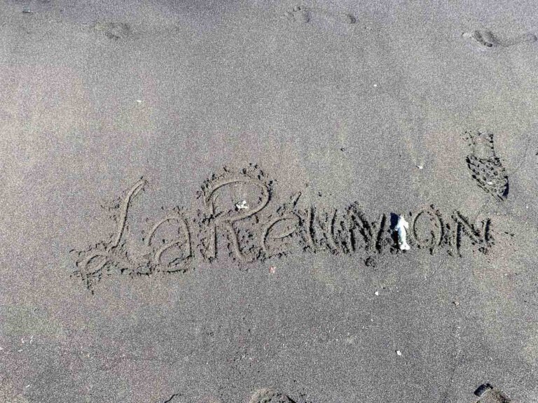 La Réunion“ in schwungvoller Handschrift in schwarzen Vulkansand gezeichnet – leicht verweht, mit Muschelrest.
