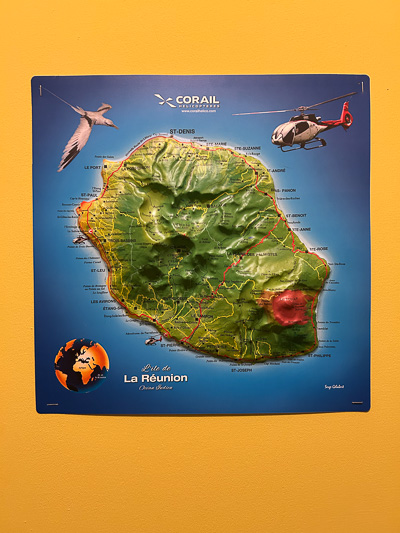Erhabene Reliefkarte von La Réunion an einer gelben Wand – mit Hubschrauber, Tropenvogel und deutlicher Topografie der Insel.