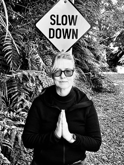 Frau mit Sonnenbrille steht unter „Slow Down“-Schild – ironisches Symbol für bewusste Reisehaltung.