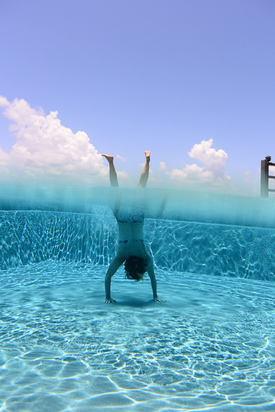 Frau im Handstand halb unter Wasser, halb über Wasser – Balance im Pool vor Himmel und Wolken