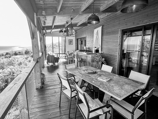 Eine überdachte Holzterrasse mit Esstisch und Stühlen, Blick ins Grüne und auf das Meer – ein halboffener Wohnbereich auf La Réunion zwischen Drinnen und Draußen.