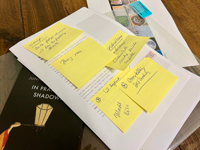 Manuskriptstapel mit mehreren gelben Haftnotizen, handschriftlich beschriftet. Darunter liegt ein Buch mit dem Titel „In Praise of Shadows“. Die Notizen wirken wie lose Gedankenbausteine – ein Turm aus Worten und Bedeutungen.