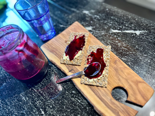Ein Glas geöffnetes Johannisbeer-Gelee auf schwarzem Steintisch, daneben zwei Knäckebrote mit dunklem Gelee und ein Löffel – einfache, ehrliche Mahlzeit.