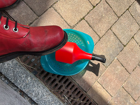 Roter Gummistiefel neben einem blauen Sieb mit roter Sprühflasche – bereit für die Johannisbeerernte am steilen Hang.