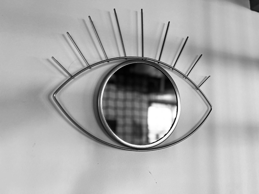 Metallischer Wandspiegel in Form eines stilisierten Auges mit langen Wimpernstreben, im Spiegel ein unscharfer Raum mit Fenster.