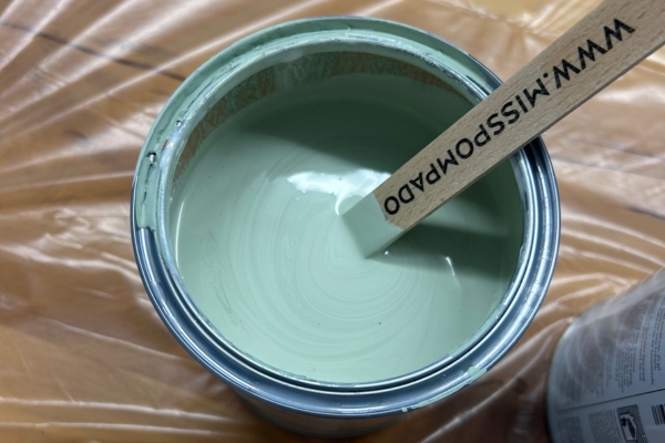 Nahaufnahme eines geöffneten Farbeimers in Salbeigrün mit Rührstab aus Holz.