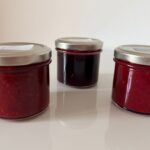 Drei kleine Marmeladengläser mit rotem Fruchtaufstrich in verschiedenen Nuancen: Kornelkirsche, Himbeere und Ribisel, ein Trio aus Sommerfarben im Glas.