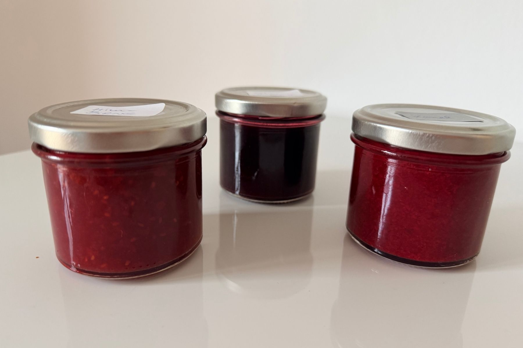Drei kleine Marmeladengläser mit rotem Fruchtaufstrich in verschiedenen Nuancen: Kornelkirsche, Himbeere und Ribisel, ein Trio aus Sommerfarben im Glas.