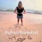 Frau steht am Strand, fotografiert ein Wort im Sand. Vor ihr der Schriftzug „Aufmerksamkeit“ – ruhig gesetzt vor der Brandung eines stillen Meeres bei Sonnenuntergang.