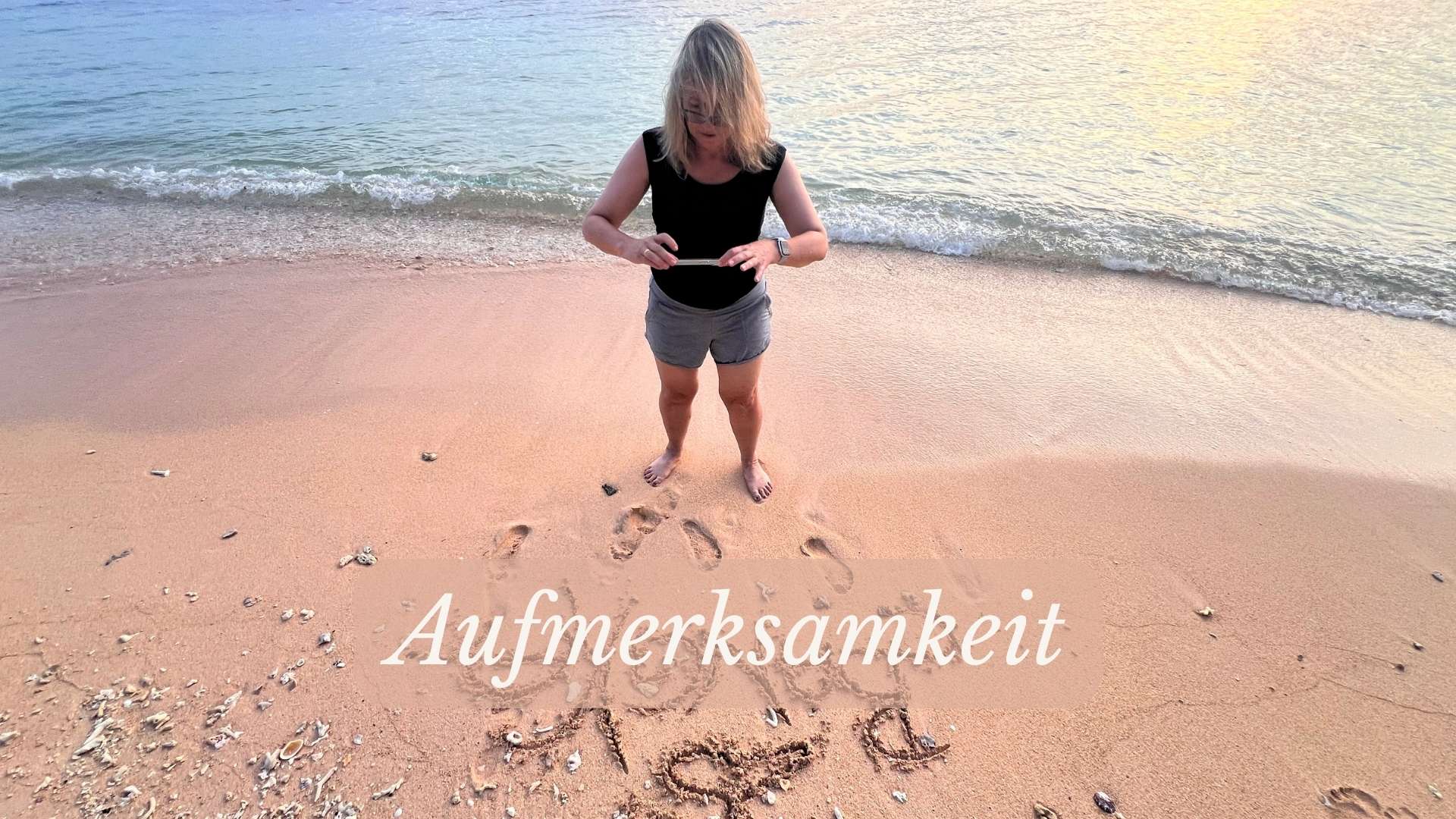 Frau steht am Strand, fotografiert ein Wort im Sand. Vor ihr der Schriftzug „Aufmerksamkeit“ – ruhig gesetzt vor der Brandung eines stillen Meeres bei Sonnenuntergang.