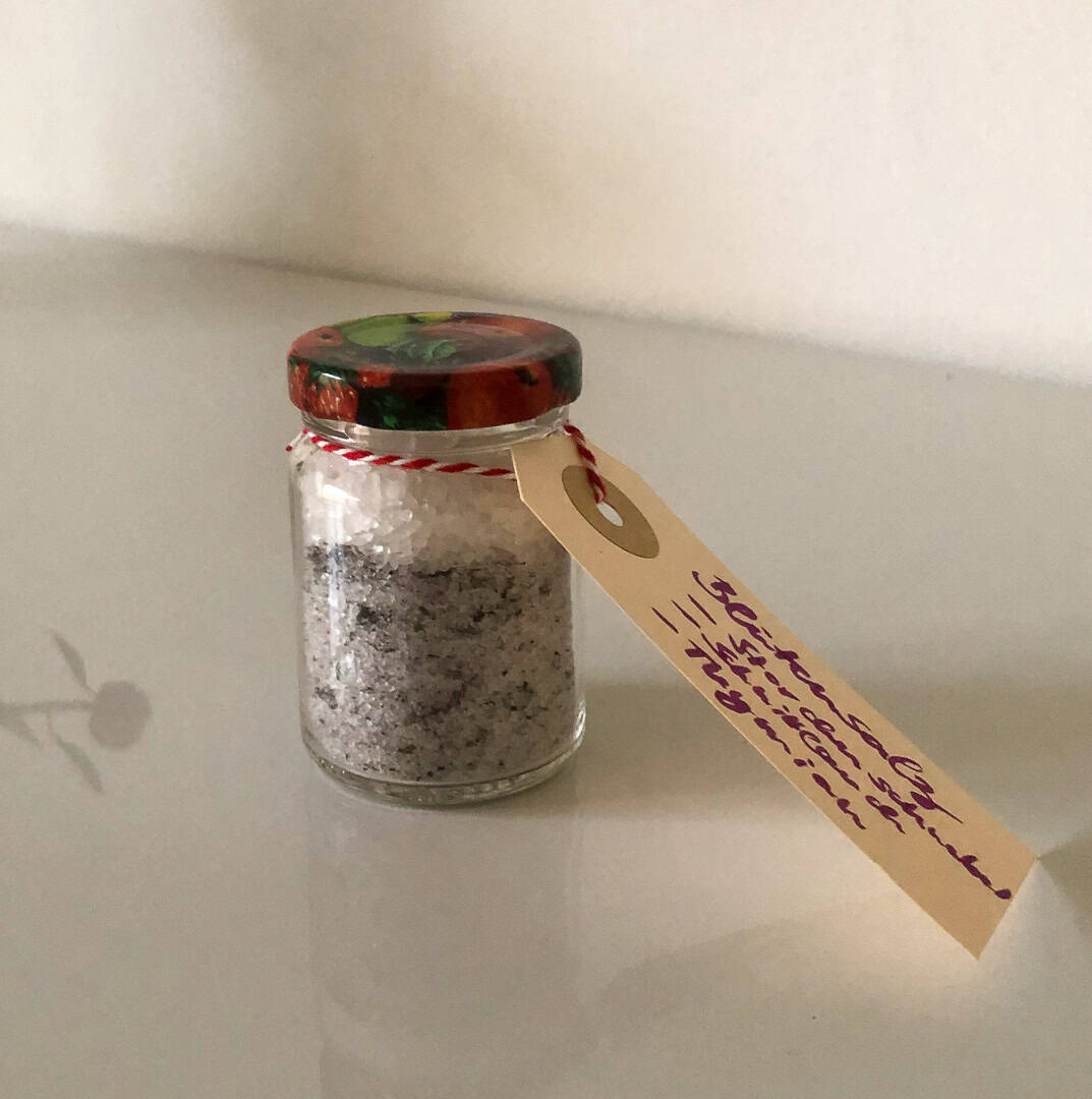 Kleines Einmachglas mit grau-violettem Blüten-Kräuter-Salz, rotem Erdbeerdeckel und handgeschriebenem Etikett an rot-weißem Bändchen.