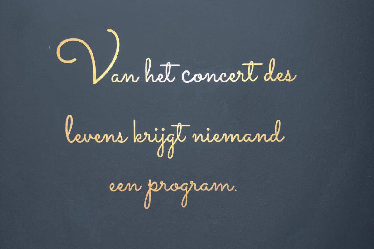 Mottospruch in gold auf dunkelbaluem Hintergrund in Schreibschrift: Van het concert des levens krijgt niemand een program