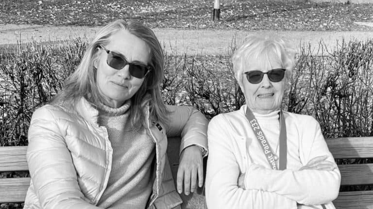 Schwarzweißfoto: Eine ältere und eine jüngere Frau sitzen nebeneinander auf einer Holzbank im Park, beide mit Sonnenbrille. Die ältere Frau trägt ein helles Schlüsselband mit der Aufschrift »petit paradis 233«.